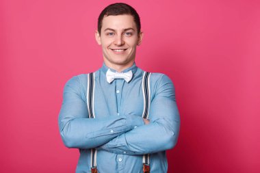 Atletik güçlü adam kollarını katlama, doğrudan kameraya bakarak, stüdyoda pembe arka plan üzerinde izole pozlar, mavi gömlek giyiyor, beyaz papyon ve soyulmuş askıları. İnsanlar ve duygular kavramı.