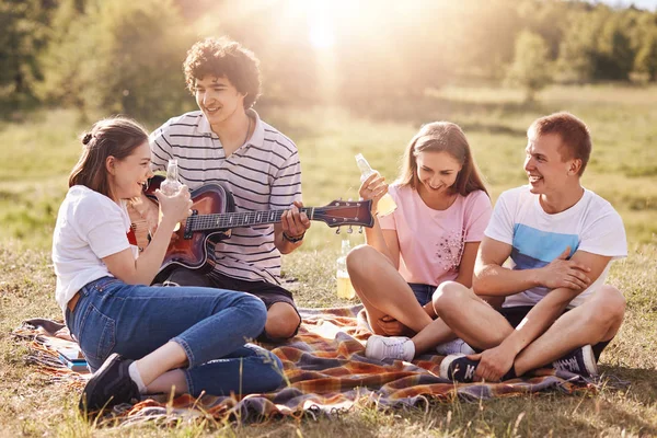 Horizontal tiro de jovens amigáveis se divertir, passar o tempo livre durante o fim de semana juntos, sentar no chão, cantar músicas para guitarra, beber bebida saborosa, desfrutar de dia ensolarado. Conceito de amizade e pessoas . — Fotografia de Stock