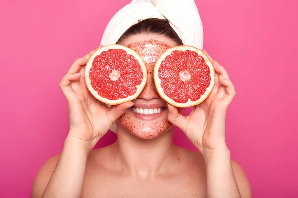 Close up portrait of positive attractive girl smiling sincerely, holding two parts of grapefruit, covering her eyes with fruite, having satisfied facial expression, being in good mood, face scrub. — Stock Photo, Image