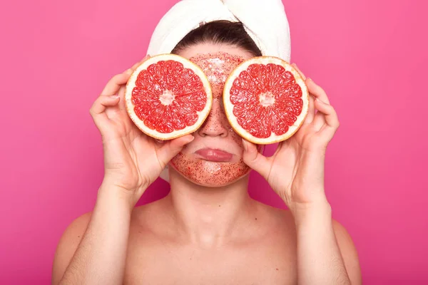Незадоволена гола жінка заморозила обличчя, тримаючи частини грейпфрута в обох руках, маючи барвистий скраб для обличчя та білий рушник. Непохитна модель пози на яскраво-рожевому фоні в студії . — стокове фото