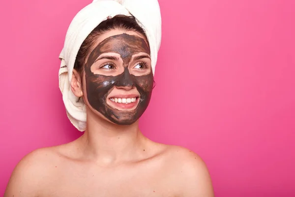 Indoor shot of young attractive woman with white towel on her head, has naked body, smilling isolated over pink background in studio, looks aside, having chocolate mask on her face. Skincare concept. — 스톡 사진