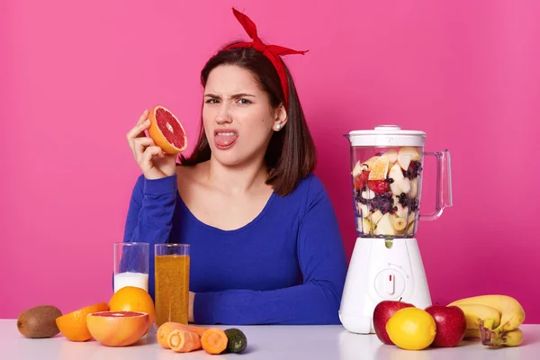 Indoor tiro de fêmea jovem descontente com cabeça vermelha brilhante na cabeça, vestindo camisola azul escuro, mostrando sua língua com nojo, segurando toranja em uma mão, parece chateado e irritado . — Fotografia de Stock