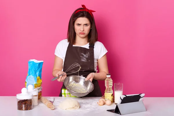 Estudio de toma de ama de casa morena decepcionada de pie en la cocina con expresión facial desagradable, sosteniendo tazón y batidor en ambas manos, no pudo mezclar todos los ingredientes de manera adecuada, cara fruncida . — Foto de Stock