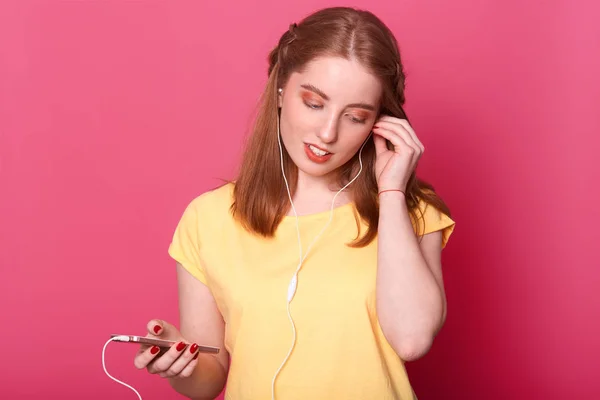 गोंडस हेअरडो आणि व्यावसायिक असलेली रोमँटिक सुंदर तरुण महिला तिचा ईयरफोन योग्य निश्चित करते, एका हातात हलका मोबाइल ठेवते. आकर्षक मॉडेल उज्ज्वल गुलाबी पार्श्वभूमीवर वेगळे पोझ . — स्टॉक फोटो, इमेज