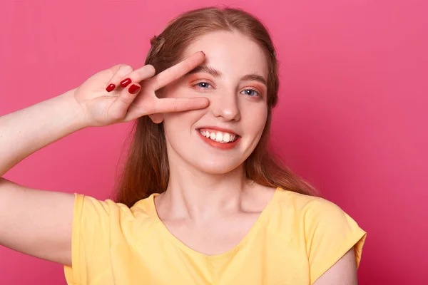 Улыбающаяся положительная модель позирует изолированным на ярко-розовом фоне в студии с победной рукой рядом с правым глазом, в жёлтой футболке, с приятным выражением лица. Свободное время и молодежная концепция . — стоковое фото