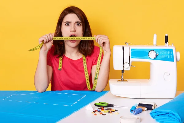 Surprised brunette seamstress holding measure tape in front of her mouth, looking directly at camera. Sewing machine, piece of blue fabric, buttons, thread situated on table. Hobby and handmade.