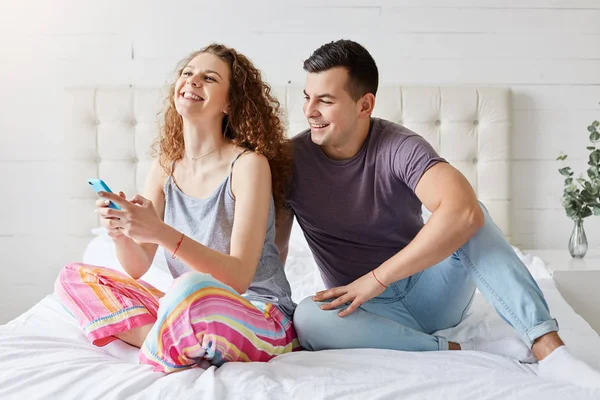 Gelukkig paar brengen ochtend tijd in de slaapkamer, het controleren van het sociale netwerk via smartphone, altijd in contact, communiceren met hun vrienden, familie met mobiele telefoons zitten lachen in comfortabel bed — Stockfoto
