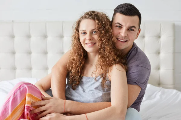 Imagem de jovem casal, cabelo encaracolado wonan e habdsome homem, sentar na cama, abraçar uns aos outros e ter uma conversa agradável, olhando diretamente para a câmera, ter expressões faciais felizes. Casal romântico apaixonado — Fotografia de Stock