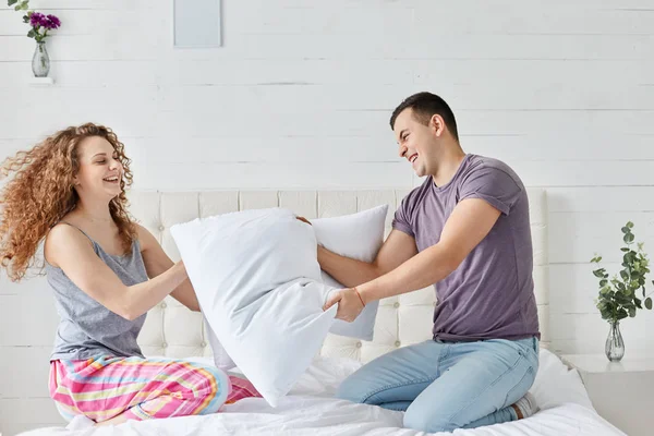 Curly woman with her attractive husband hold big pillow, have fun in bed in morning, romantic couple spend time together at home at weekend, young family enjoying spare time with each other. — Stock Photo, Image