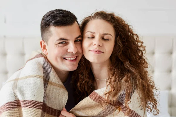 Retrato de casal feliz bonito sentado na cama abraçar uns aos outros. Menina bonita com cabelos longos encaracolados senta-se com os olhos fechados com o marido, cara abraça sua namorada sob xadrez quente no quarto . — Fotografia de Stock