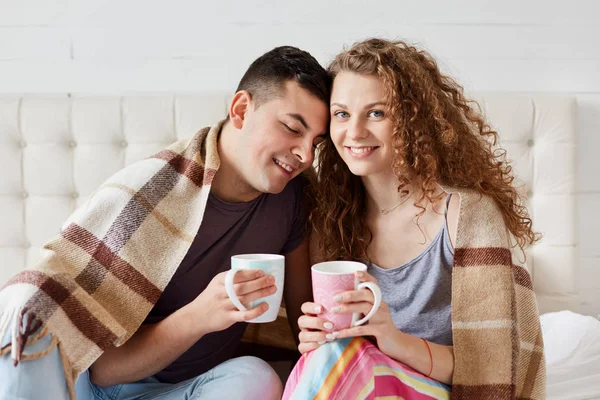 Imagem de jovem casal feliz beber café ou chá na cama de manhã, sentado sob xadrez quente, desfrutando de fim de semana, mulher olha para a câmera, tem expressão facial agradável, homem inclinado para a namorada . — Fotografia de Stock