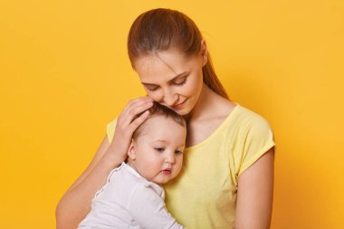 Sevgi ile onun bebek kız sarılma güzel mutlu anne yakın portre, anne at kuyruğu ile rahat t bok giyer, sarı arka plan üzerinde izole fotoğraf stüdyosunda poz. Annelik ve aile kavramı.