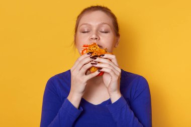 Pasta parçası tutan ve onu koklar, kapalı gözlü pasta yemek, mavi gömlek giyer, lezzetli ev yapımı pasta sahiptir güzel kadın portresi kadar yakın. Genç kadın model sarı stüdyo duvar üzerinde pozlar.