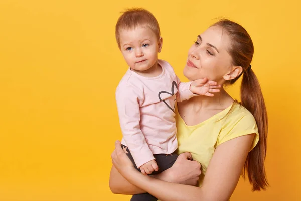 Studio shot of loving mother holding her baby girl, toddler looking aside, mom admires her charming doughter isolated over yellow studio wall. Motherhood, happyness, children concept. Copy space. — Stock Photo, Image