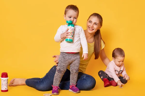 Image of loving attractive mother looks after her little children, twins playing with mommy. Playful kids drink tasty baverage from her bootle while her sister eating coockies. Infants are hungry. — Stock Photo, Image