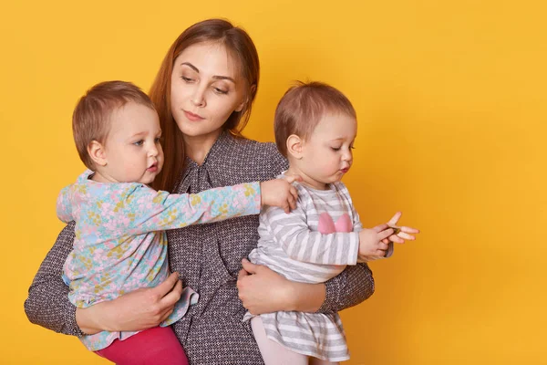Portrait of responsible attentive mother spending time with her little children, holding twin girls in arms, listening to them seriously. Cute kid rises her arm, showing direction, looks curious. — Stock Photo, Image