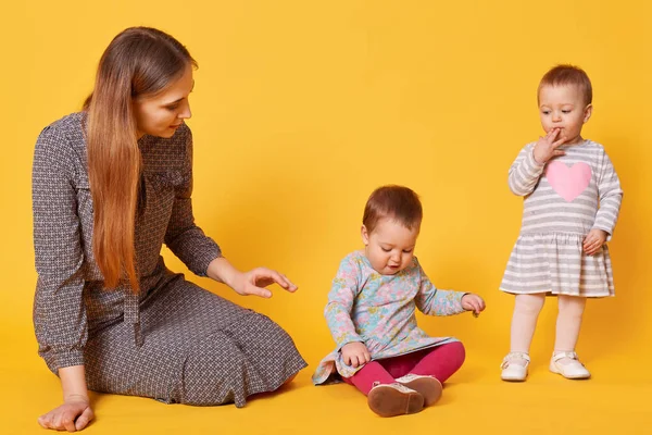 Young adorable caring mother looks after her little kids, sitting on floor with one of twin girls. Sweet child puts one hand on her mouth, looking attentively at her sister. Love and family concept. — Stock Photo, Image