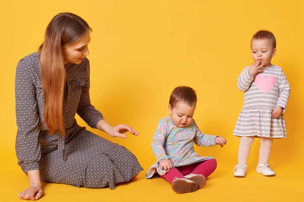 Joven madre cariñosa adorable cuida de sus hijos pequeños, sentado en el suelo con una de las niñas gemelas. Dulce niña pone una mano en su boca, mirando atentamente a su hermana. Amor y concepto familiar . — Foto de Stock