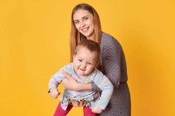 Smiling positive young lady spends time with her kid, holding in hands, playing with her, making her laugh. Fair haired little girl enjoys game with her caring mom. Family activities concept. — Stock Photo, Image