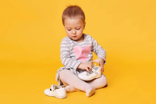 Menina bonita de cabelos bonitos vestindo vestido despojado com poses de coração isolado sobre fundo amarelo brilhante. Doce criança magnética senta-se no chão no estúdio segurando e observando seus sapatos brancos . — Fotografia de Stock