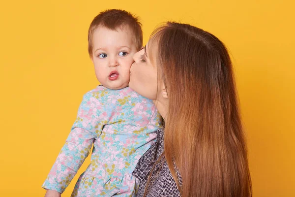 Indoor shot of beautiful tender woman holding her daughter, kissing her in cheek. Tiny cute little girl looks aside, opens her mouth with interest, wearing light blue dress. Family and care concept. — Stock Photo, Image