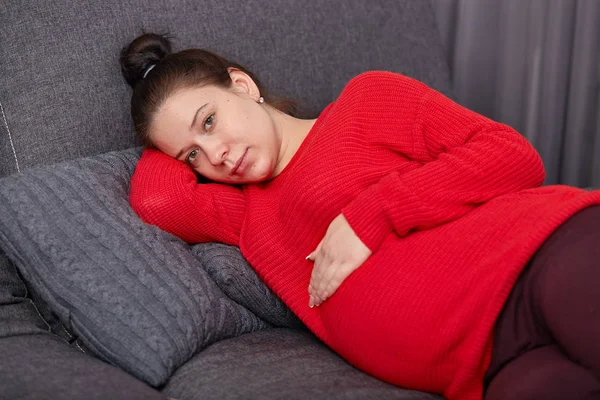 Image of sleepy tired pregnant lady taking nap, keeps hands on belly, dressed red sweater, enjoying calm atmosphere, lies on grey comfortable sofa in cosy woom. Pregnancy, motherhood concept. — Stock Photo, Image