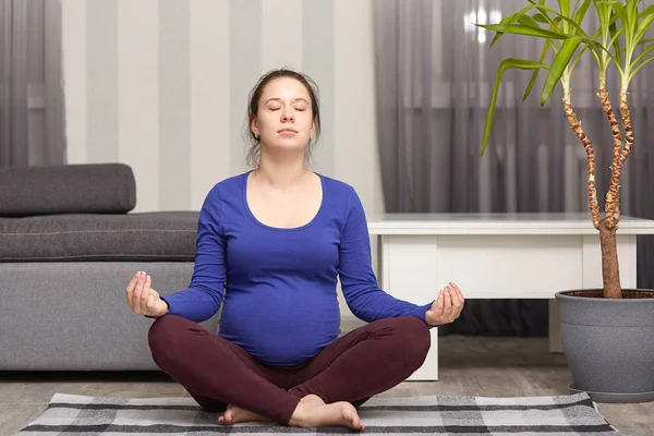 Foto de mujer morena con manojo en la cabeza, espera un hijo, hacer ejercicio en casa, practicar yoga y meditación, posa en apartamento moderno, viste suéter azul y polainas, maternidad, embarazo deportivo . — Foto de Stock