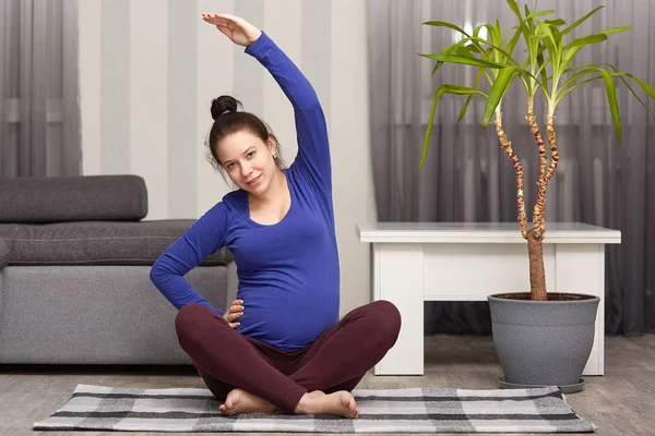 Image of charming woman expects child, does sport at home, takes care of her healthy, wears blue sweater and leggings, has bunch on head, raises one hand, has healthy lifestyle during pregnancy. — Stock Photo, Image