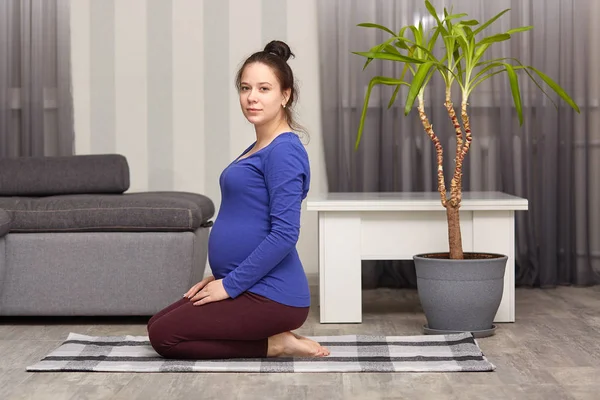 Side view of brunette woman expects child, exercising at home, practices yoga, poses in modern apartment, wears blue sweater and leggings, looks directly at camera, sits on floor. Motherhood concept. — Stock Photo, Image