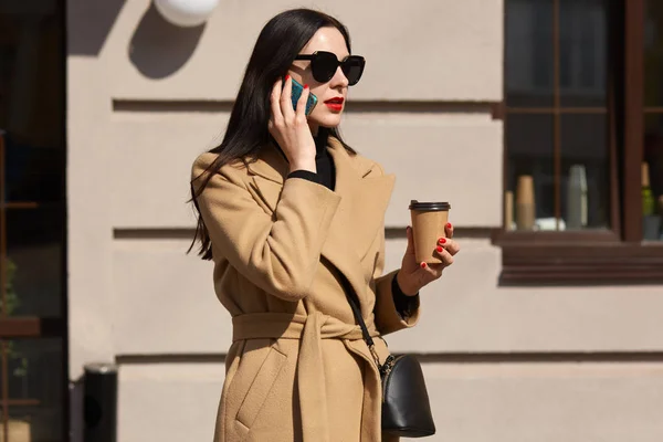 Retrato de una atractiva mujer ocupada usando abrigo beige y sosteniendo café para llevar mientras habla por teléfono, discutiendo asuntos importantes. Joven morena mujer de negocios mira a un lado, dejando café . — Foto de Stock