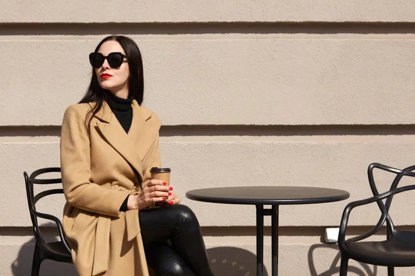 Charmig Snygg modell med rött läppstift bär stora trendiga solglasögon, beige päls, svart tröja och byxor, hålla papercup av starkt kaffe, sittande utomhus över ljus vägg, njuter solljus. — Stockfoto