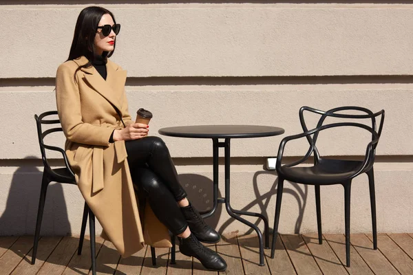 Fashionable trendy female sits on chair near light coloured building, waiting for someone on terrace with coffee in her hand, looking aside, enjoying her free time alone during warm spring day. — Stock Photo, Image