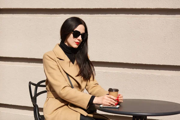 Närbild porträtt av vackra snygg modell sitter vid brunt träbord, dricka kaffe, ser lugn. Fashionabla ung dam bär beige trendig päls, svarta accessoarer och tröja, röd manikyr. — Stockfoto