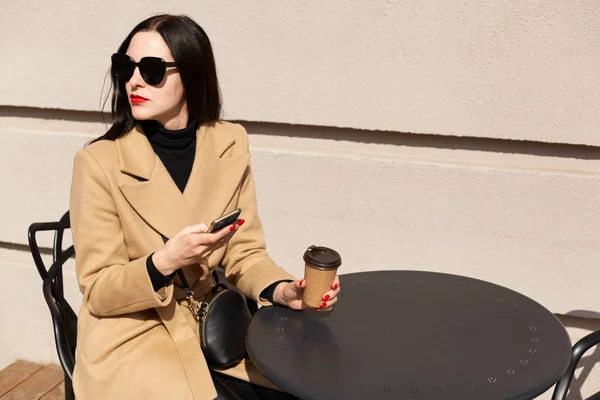 Primer plano retrato de mujer elegante atractiva sentada sola con una taza de café fuerte y su teléfono móvil, viendo lo que está sucediendo a su alrededor, distraído por el ruido de la ciudad. Concepto de moda . — Foto de Stock