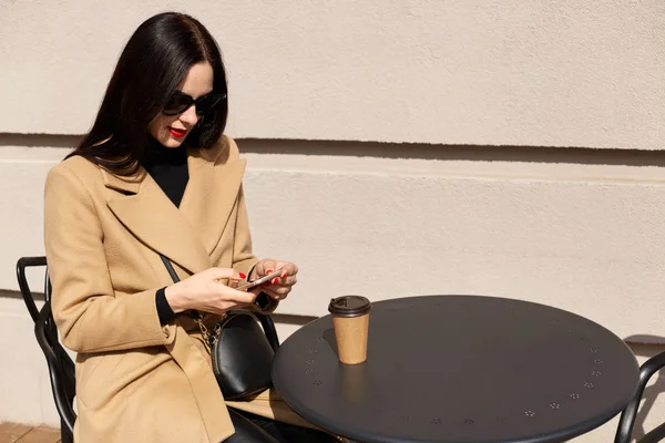 Primer plano retrato de hermosa mujer joven elegante con abrigo beige y beber café para llevar mientras almorza. Bonita morena revisando sus sitios de redes sociales, charlando . — Foto de Stock