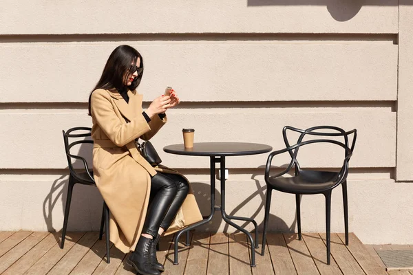Esbelta hembra de pelo negro se sienta a la mesa al aire libre, tomando una taza de bebida caliente, relajándose durante el tiempo libre, tomando fotos de escenas urbanas en el cálido y soleado día de primavera. Concepto de personas y espacio urbano . — Foto de Stock