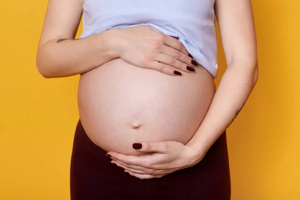 Mujer embarazada joven sin rostro sostiene su gran vientre con la mano aislada sobre el fondo amarillo. Modelo embarazada siendo fotografiada en un estudio fotográfico. Futura madre espera bebé y posando con abdomen desnudo . — Foto de Stock