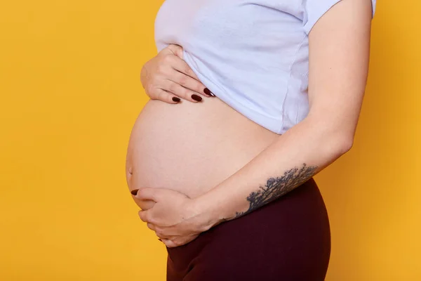 Side view of pregnant female with naked belly isolated over yellow background. Woman with tattoo on hend dressed casually being photographed in photo studio. Motherhood and pragnancy concept. — Stock Photo, Image