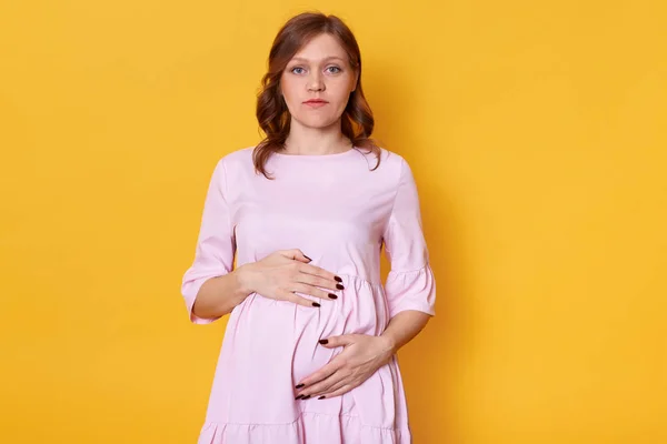 Close up portrait of adorable pregnant female in gentle dress, keeps hands on her belly, touching her baby, wearing pink dress, posing isplated over yellow background in studio. Motherhood concept. — Stock Photo, Image