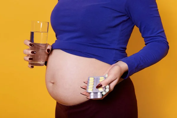 Mujer embarazada sin rostro joven sosteniendo un vaso de agua y pastillas, de pie aislado sobre fondo amarillo, modelo fotografiado con el abdomen desnudo. La futura madre espera un bebé. Concepto de embarazo . — Foto de Stock