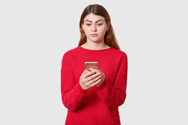 Retrato de mujer linda confundida con cabello liso oscuro, usa suéter rojo, sostiene el teléfono móvil en la mano, mirando a su pantalla, aislado sobre fondo de estudio blanco. Concepto de amoción de personas . — Foto de Stock