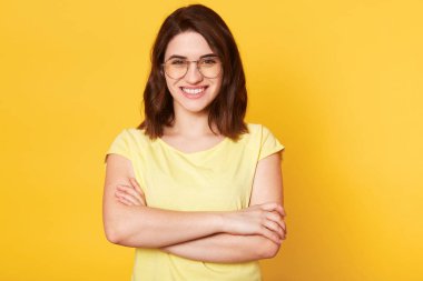 Sarı stüdyo arka plan üzerinde izole katlanmış elleri ile güzel gülümseyen kadın Portresi, rahat giyinmiş, yuvarlak gözlük vardır, mutlu yüz ifadesi vardır, iyi bir ruh hali içinde olmak, harika bir haber var.