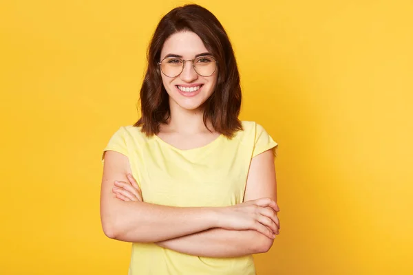 Ritratto di bella donna sorridente con le mani piegate isolato su sfondo studio giallo, vestito con disinvoltura, ha occhiali rotondi, ha espressione del viso felice, essendo di buon umore, ha grandi notizie . — Foto Stock