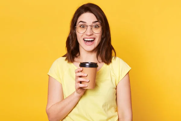 Retrato de cerca de la joven aislada en el fondo del estudio amarillo, posando con expresión facial feliz mientras bebe café tekeaway, tiene sentimientos positivos y sonrisa dental, se para con la boca abierta — Foto de Stock