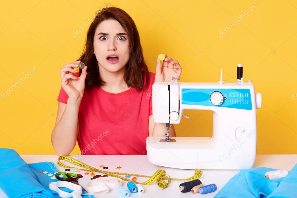 Close up portrait of brunette woman in sewing workshop, sitting with opend mouth, has astonished facial expression, holding buttons in hands, surraunded with sewing equipment. Design concept.