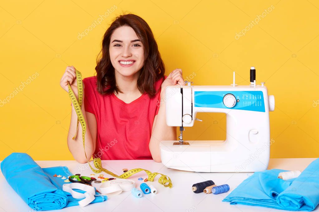 Close up portrait of charming woman seamstress sitting at table with sewing machine on yellow background in studio, dressmaker sews new dress, looks happy, holds measure tape, designer makes outfit.