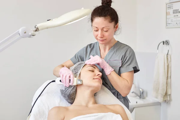 Beeld van brunette cosmetologist draagt grijze kleren doen Ultra Sonic procedures voor jonge dame liggend op couh in cosmetologie kliniek, houdt de ogen gesloten, het dragen van de wegwerp dop en verpakt witte handdoek. — Stockfoto