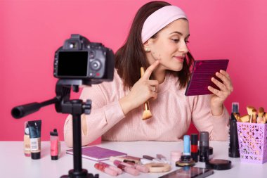 Yüzünde akne ile üzgün kadın blogger yakın portre, kozmetik fırça tutar, aynaya bakar, pembe arka plan üzerinde izole gül gömlek ve saç bandı giyer. Online çeviri, kayıt videosu