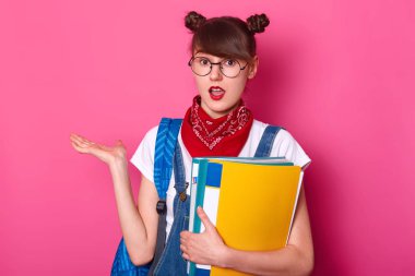 İki demet ile öğrenci portresi, t shirt giyer, tulum, bandana, kağıt klasör tutar, bir kenara palmiye alır, yüz ifadesi şaşırttı, pembe arka plan üzerinde izole açık ağız ile pozlar.