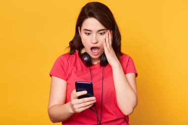 Şaşırmış genç Avrupalı esmer kadın Stüdyo çekim şok ifade ile yanak elini tutar, tutarak ve akıllı telefon bakarak, yüzünde sürpriz tepki, kırmızı t sirt giyen, koyu saçlı.